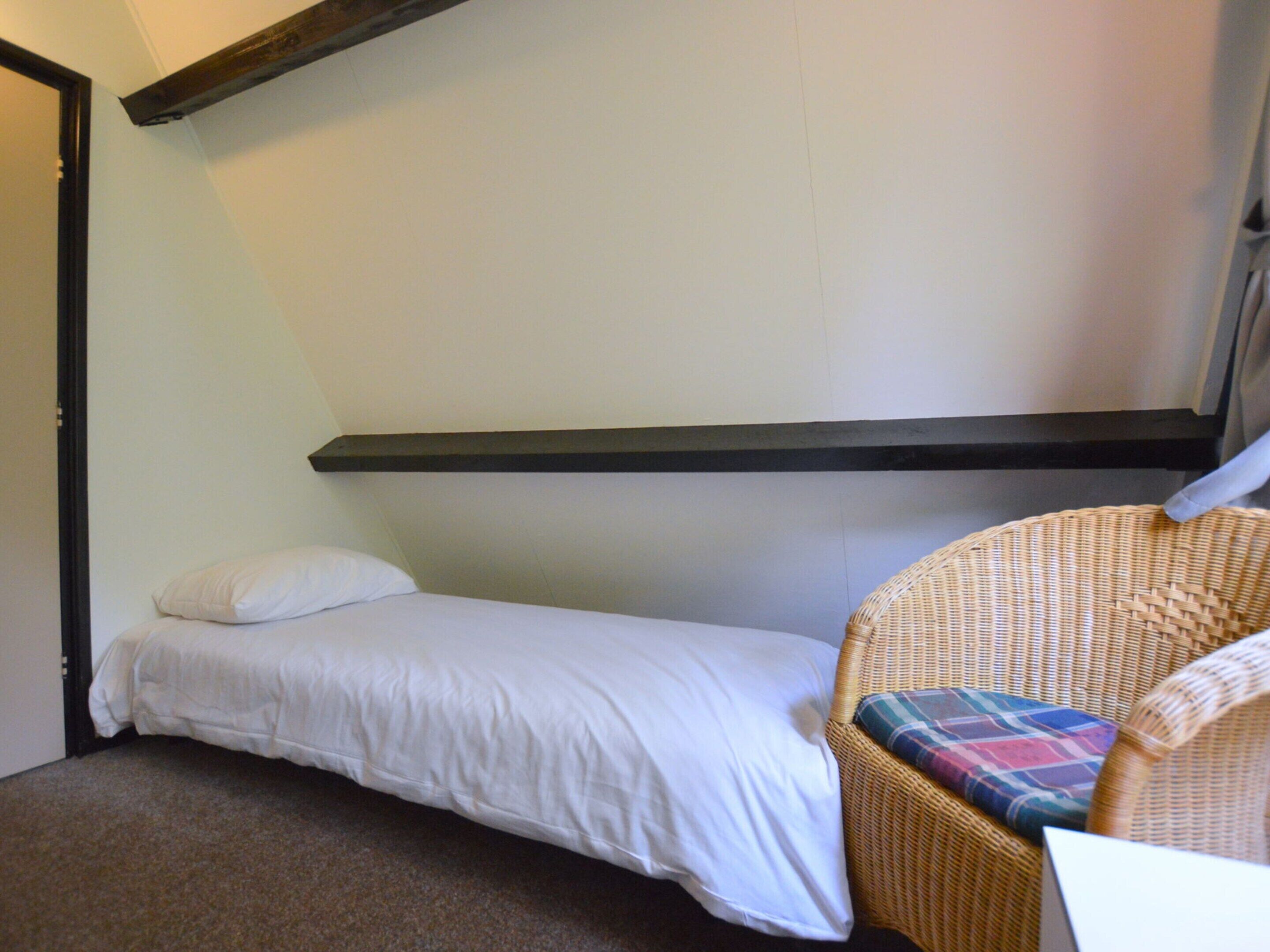 Une petite chambre avec un lit simple, un lit sans fioritures, une chaise en osier agrémentée d’un coussin coloré et un mur sobre.