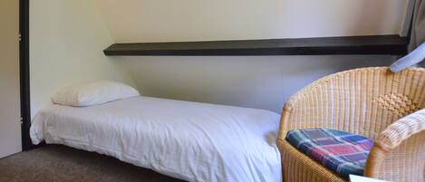 Une petite chambre avec un lit simple, un lit sans fioritures, une chaise en osier agrémentée d’un coussin coloré et un mur sobre.