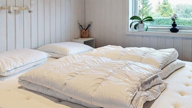 Un letto con un piumone bianco trapuntato e cuscini, un comodino con una pianta e un oggetto decorativo, e una finestra con vista sul verde.