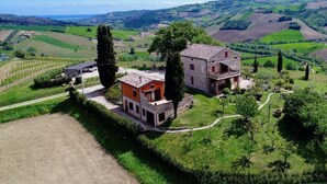 A two-story stone house with a balcony, surrounded by lush green fields and vineyards, overlooking rolling hills. - Cypressa Modern Retreat (Montefiore dell'Aso)