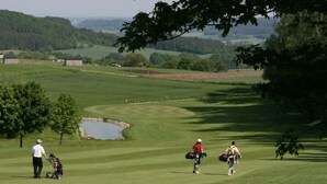 Un campo de golf con un estanque, árboles y colinas al fondo.