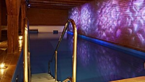 Un área de piscina con una escalera metálica que baja al agua, rodeada de vigas de madera y una pared iluminada en morado.