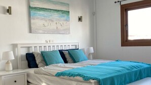 A bedroom with a white bed, teal bedding, a bench, a nightstand, a lamp, and a painting of a beach scene. - Mesmerising Apartment in Playa Blanca (Yaiza)