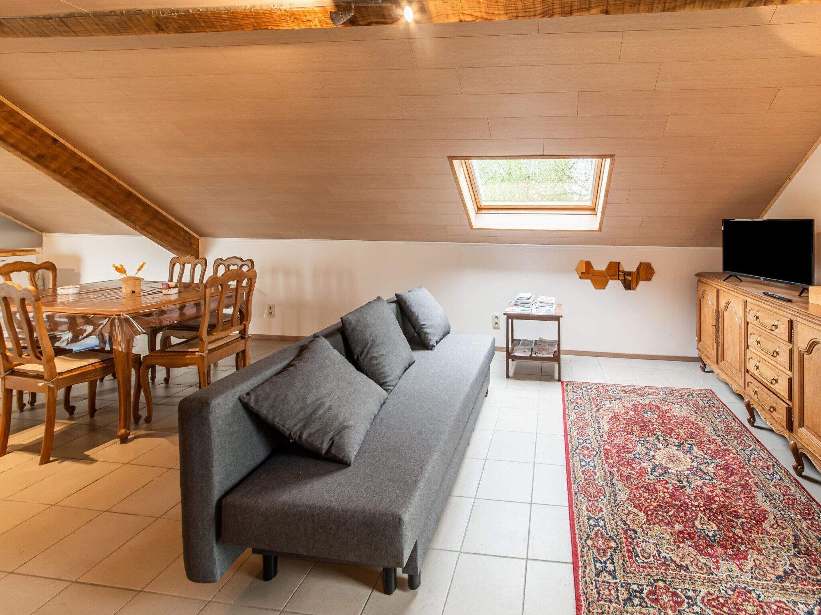 A cozy attic bedroom with a skylight, a gray sofa, a wooden dining table with chairs, a TV on a wooden cabinet, and a patterned rug.