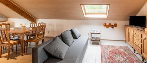 A cozy attic bedroom with a skylight, a gray sofa, a wooden dining table with chairs, a TV on a wooden cabinet, and a patterned rug.