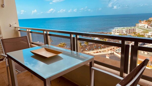 Un balcón con una mesa y sillas de vidrio, con vistas a la costa, edificios y el mar.