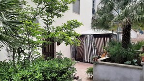 A courtyard with a variety of plants, a palm tree, and a striped awning. - Apartment in Matera Near Historic Center (Matera)