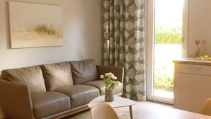 Ein Wohnzimmer mit einem braunen Ledersofa, einem kleinen runden Tisch mit Blumenvase und einem Fenster mit gemusterten Vorhängen.