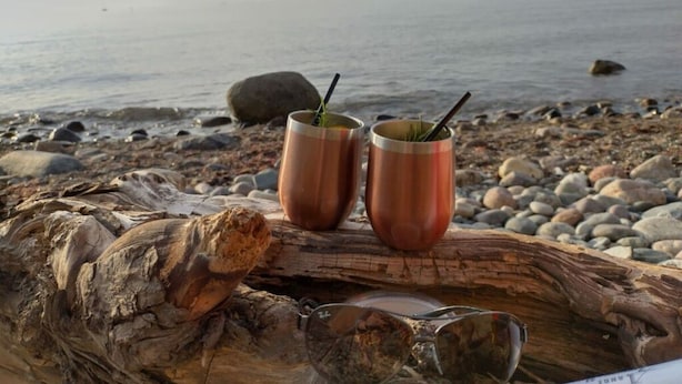 Dos tazas de cobre sobre un tronco, con un par de gafas de sol a un lado, sobre una playa rocosa con el mar al fondo.