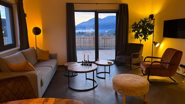 Una sala de estar acogedora con sofá, reposapiés y sillones, una mesa de centro de madera, una lámpara de pie y vistas a montañas a través de grandes ventanales.