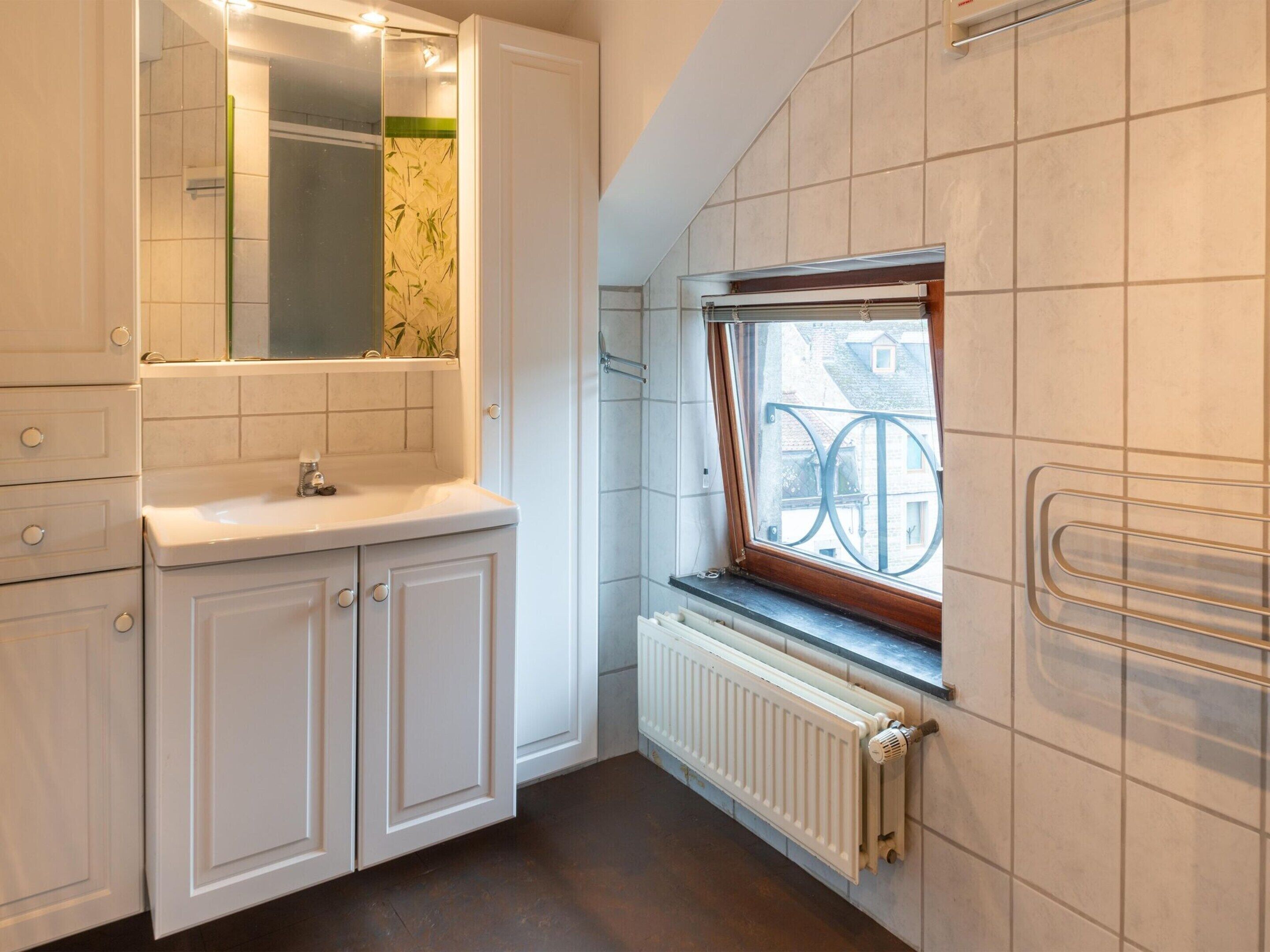 A bathroom with a white sink, mirror, and cabinets, a window with a view of buildings, and a radiator.