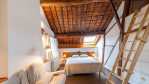 Una habitación estrecha con techo de madera, una cama, una silla y una escalera.