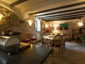 A rustic dining area with stone walls, wooden beams, and a stone fireplace. There is a dining table with chairs, a kitchen area with a blender, and a television. - House of Olives Comfortable Holiday Residence (Dobrinj)