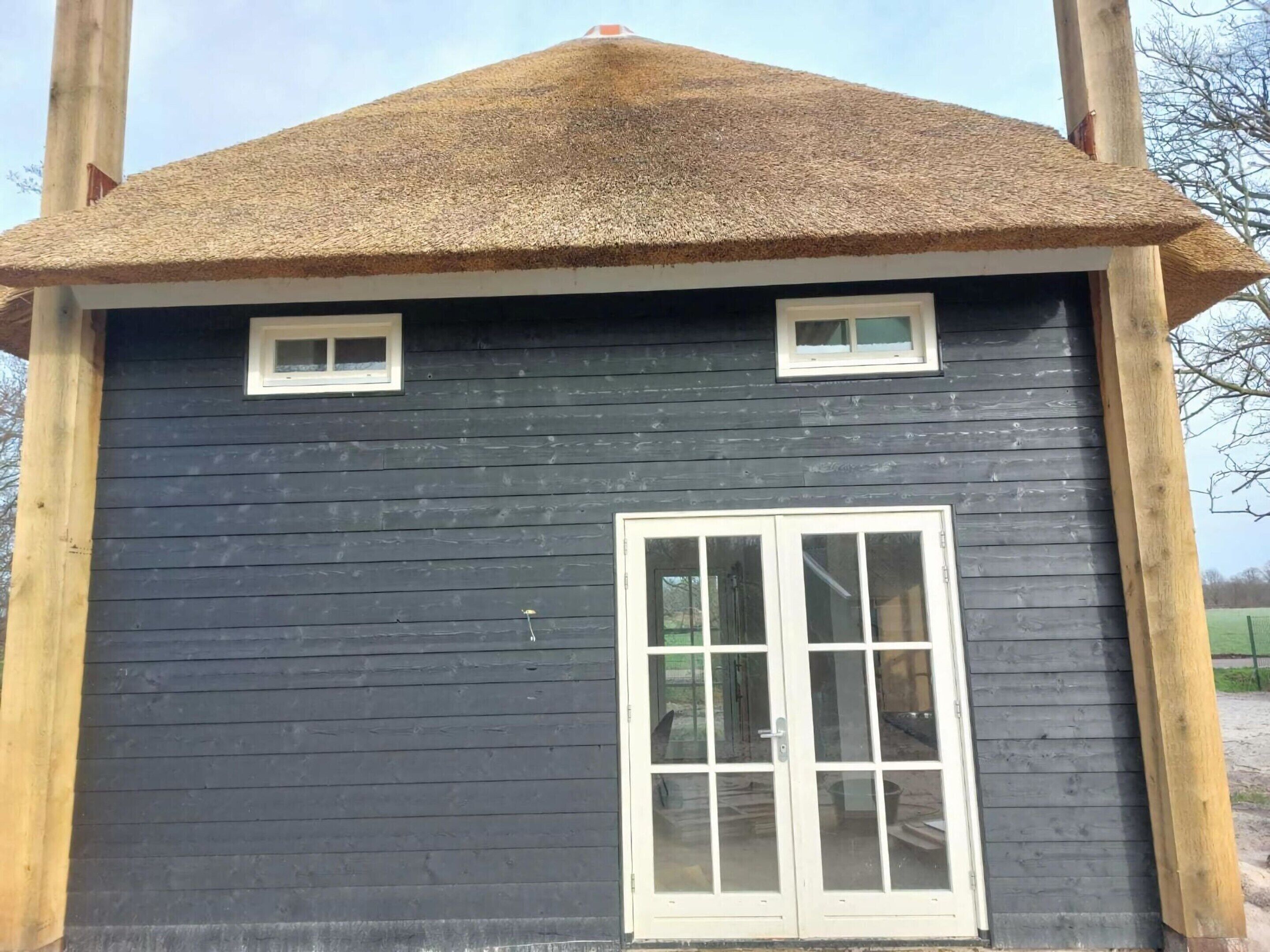 A small, dark wooden building with a thatched roof, a single large window, and two small windows.