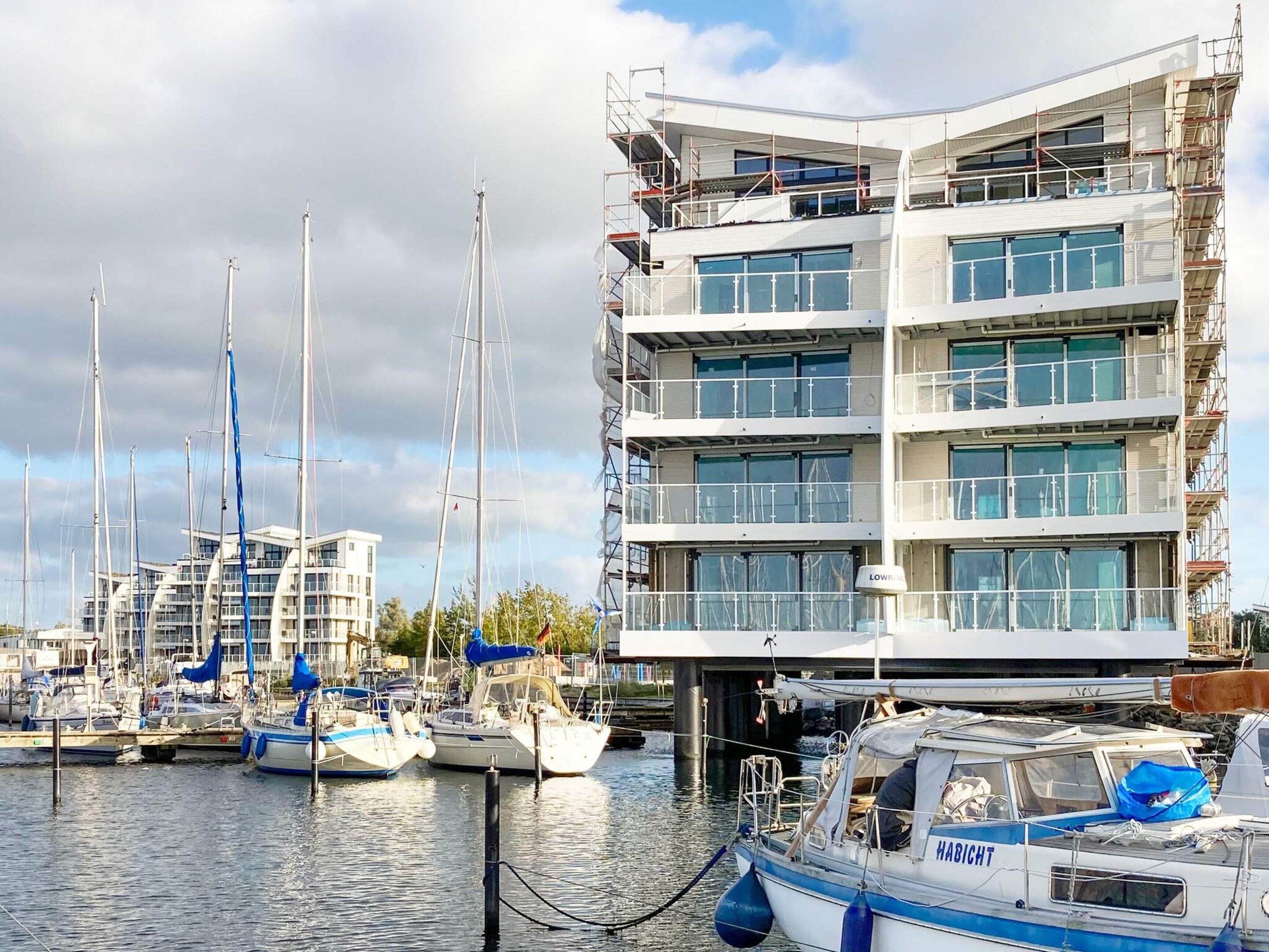 A marina with multiple boats docked, a modern multi-story building with balconies, and a clear sky.