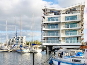 A marina with multiple boats docked, a modern multi-story building with balconies, and a clear sky.
