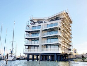 Un edificio de varios pisos con balcones, situado sobre el agua y con barcos al fondo.