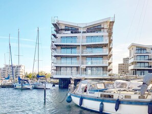 Un edificio de varios pisos con balcones, un puerto deportivo con barcos y un cielo despejado.