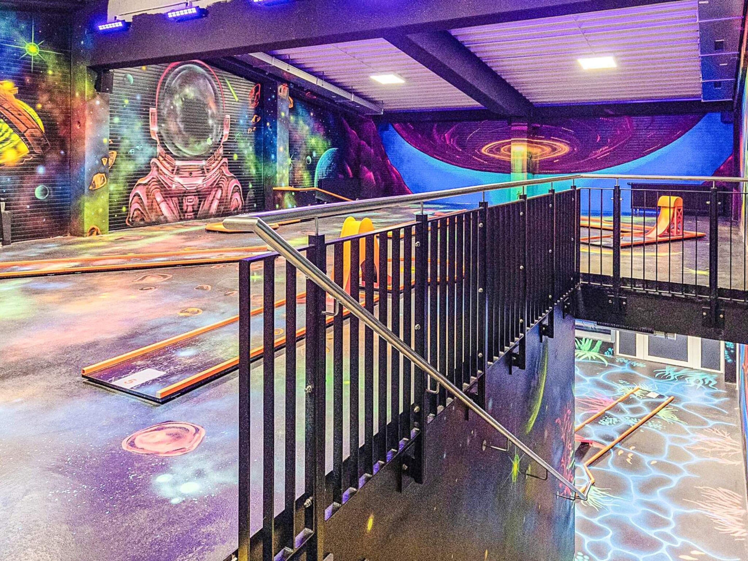 Un área temática del espacio con un mural de un astronauta y planetas, una escalera con barandillas metálicas y una zona de juegos con un tobogán.