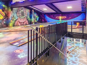 Área temática espacial com um mural de astronauta e planetas, escada com corrimão de metal e área de recreação com escorrega.
