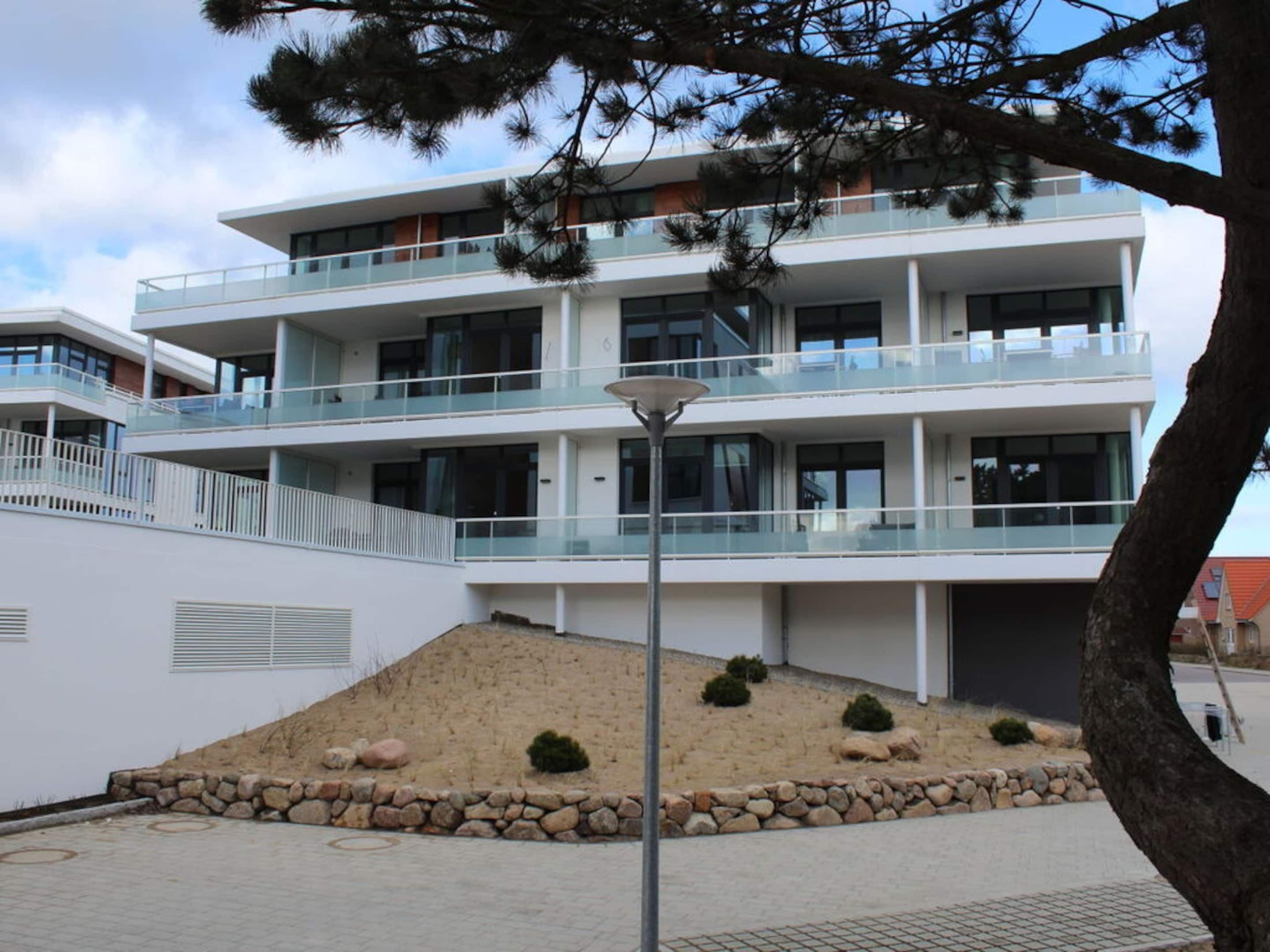 Een modern meerlaags gebouw met balkons, een aangelegde omgeving met rotsen en struiken, en een heldere lucht.