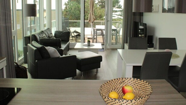 A modern living room with a sofa, a dining table, and a kitchen area. There is a balcony with outdoor furniture visible through the glass doors. - Seaside, South Cape Apartment D5 (Neustadt in Holstein)