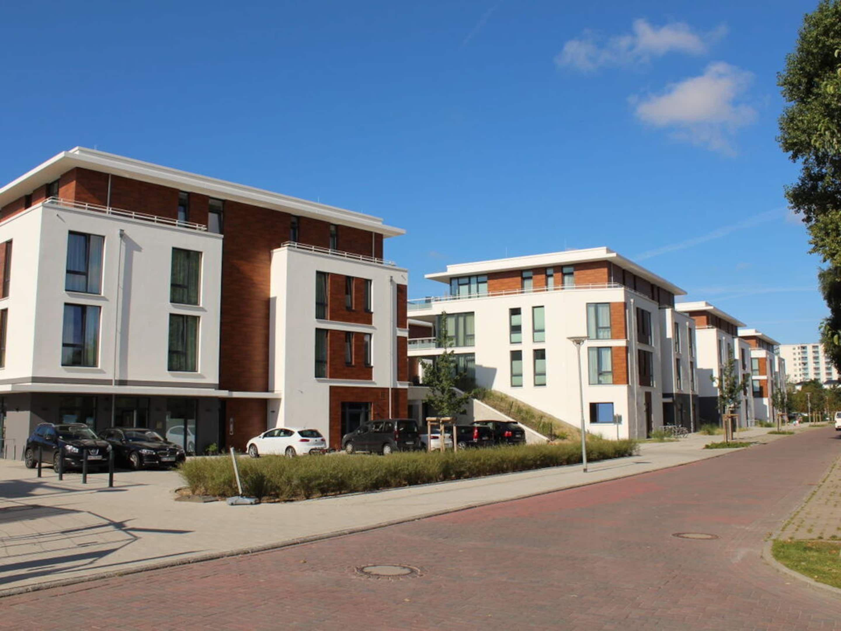 A modern apartment building with a brick facade, multiple balconies, and a paved street with parked cars.