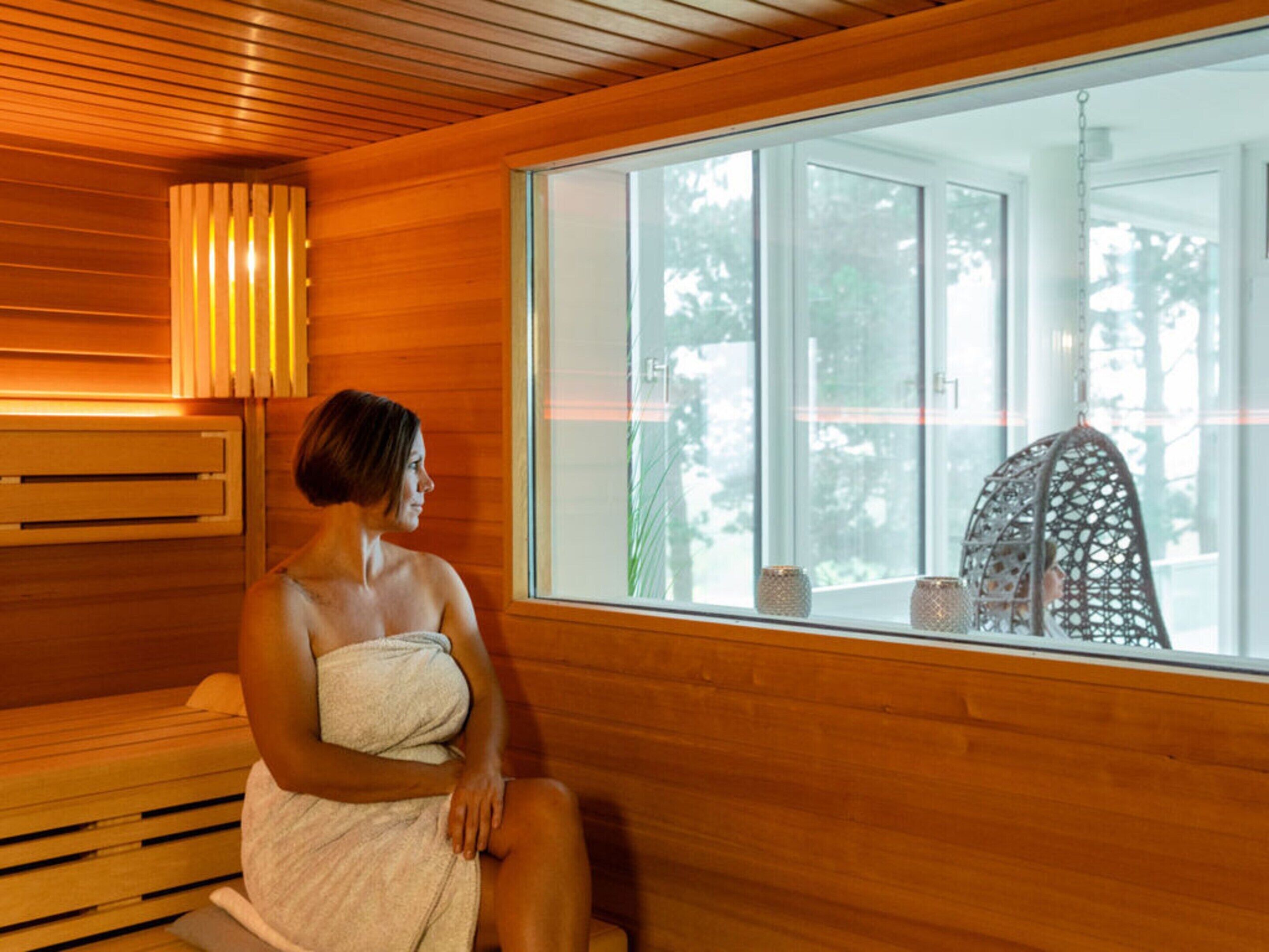 A person sitting on a bench in a wooden room with a large window, a hanging chair, and a wall-mounted lamp.