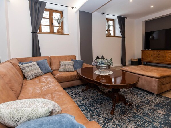 Un salon avec un canapé en cuir marron, un pouf assorti, une table basse en bois surmontée d’un vase décoratif, un grand téléviseur à écran plat et un tapis à motifs.