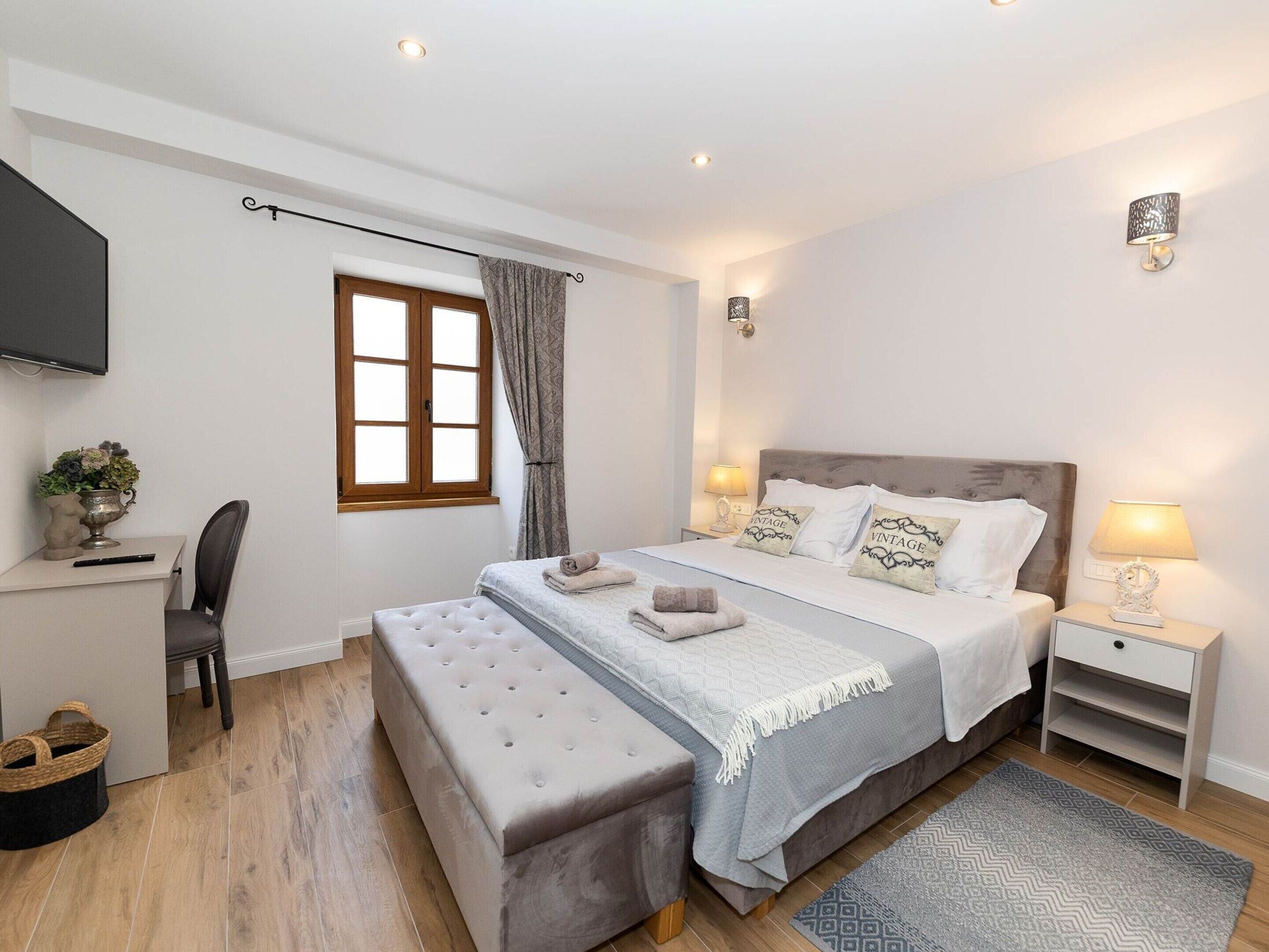 Une chambre à coucher comprenant un lit, des tables de chevet, un téléviseur, un bureau avec une chaise, une fenêtre avec des rideaux et une lampe fixée au mur.