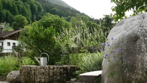 A stone bench with a metal watering can, surrounded by lush greenery and large rocks. - Berg-loft Ramsau (Ramsau bei Berchtesgaden)