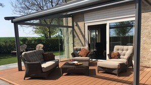 Une terrasse extérieure aménagée avec des meubles en osier, une véranda vitrée et une vue sur un jardin.