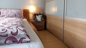 Una cama bien hecha con un edredón estampado, una lámpara de noche y una mesita con una planta.