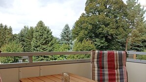 Balcón con una mesa de madera y una silla a rayas, con vistas a un área boscosa.