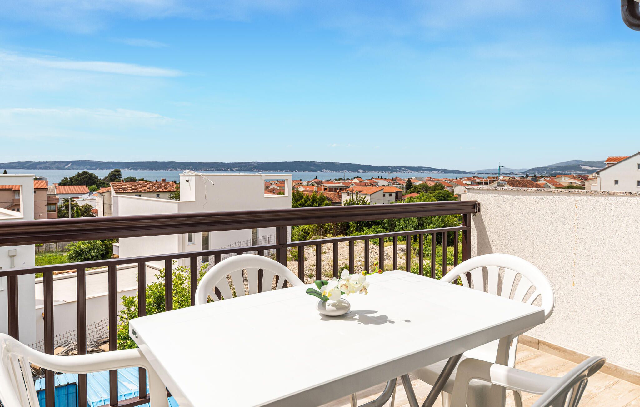 Ein Balkon mit Tisch und Stühlen, mit Blick auf eine Küstenstadt mit roten Dächern und blauem Himmel.