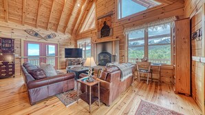 A cozy cabin interior with a wooden ceiling, a fireplace, a sofa, a television, and a desk.