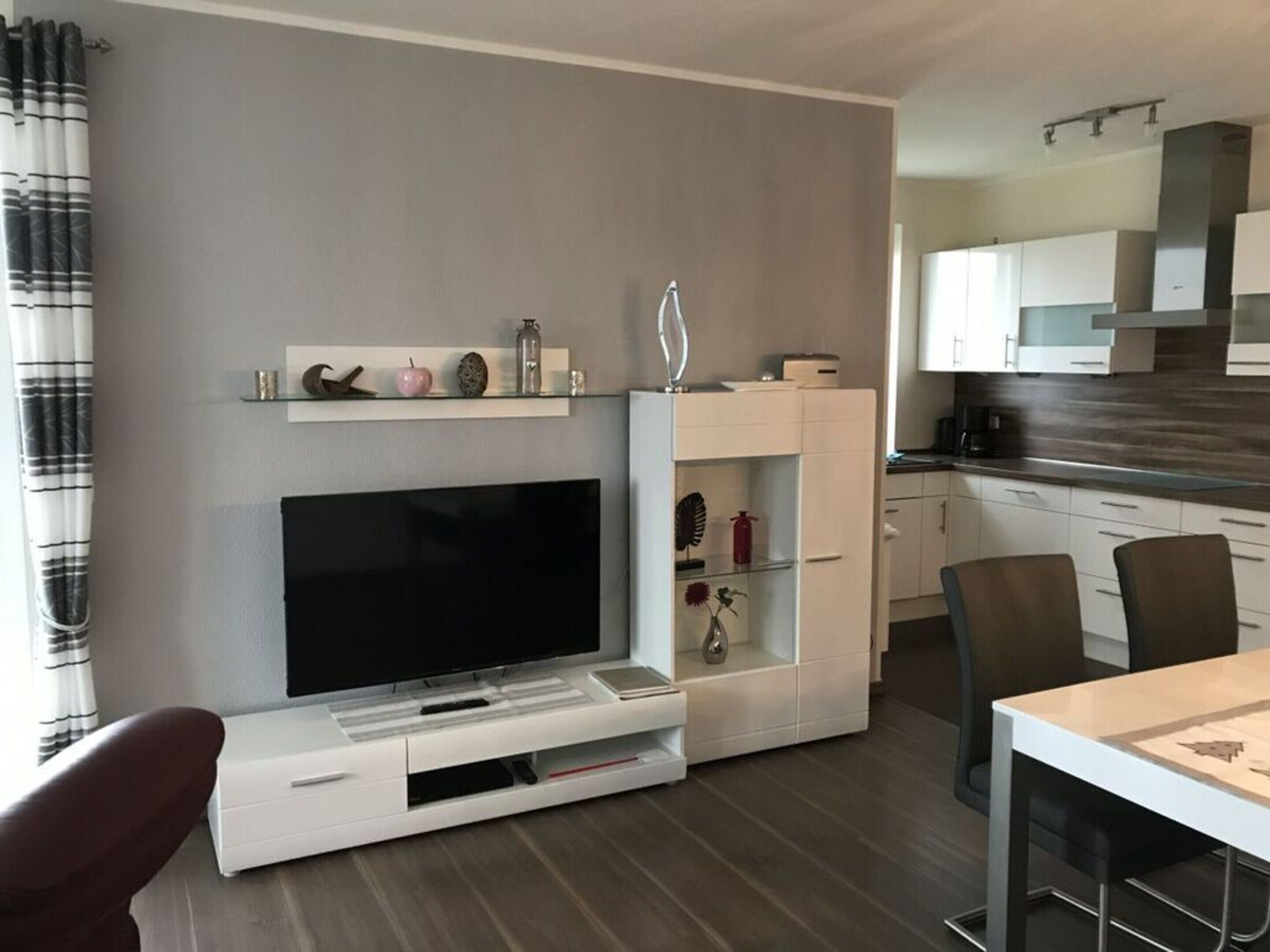 A modern living room with a flat-screen TV, a white entertainment unit, and a dining area with a table and chairs.