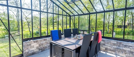 A glass-enclosed patio with a table and chairs, a brick wall, and a view of greenery.