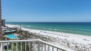 Une vue sur la plage depuis un balcon, avec une piscine, des parasols de plage et un ciel dégagé.