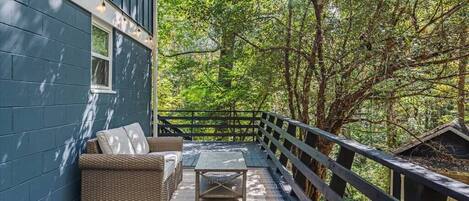 A covered outdoor area with a sofa and table, surrounded by a wooden deck and trees.