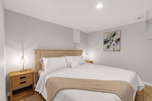 A bedroom with a wooden headboard, a bed with white bedding, a nightstand with a lamp, and a wall-mounted artwork.