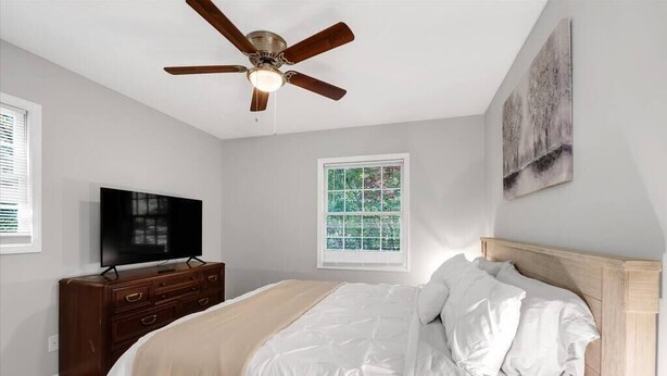 Un dormitorio con cama, ventilador de techo, televisor, cómoda y ventana con persianas.