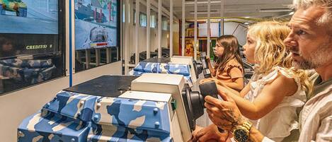 A room with a large screen displaying a video game, a table with a camouflage-patterned box, and a person holding a game controller.