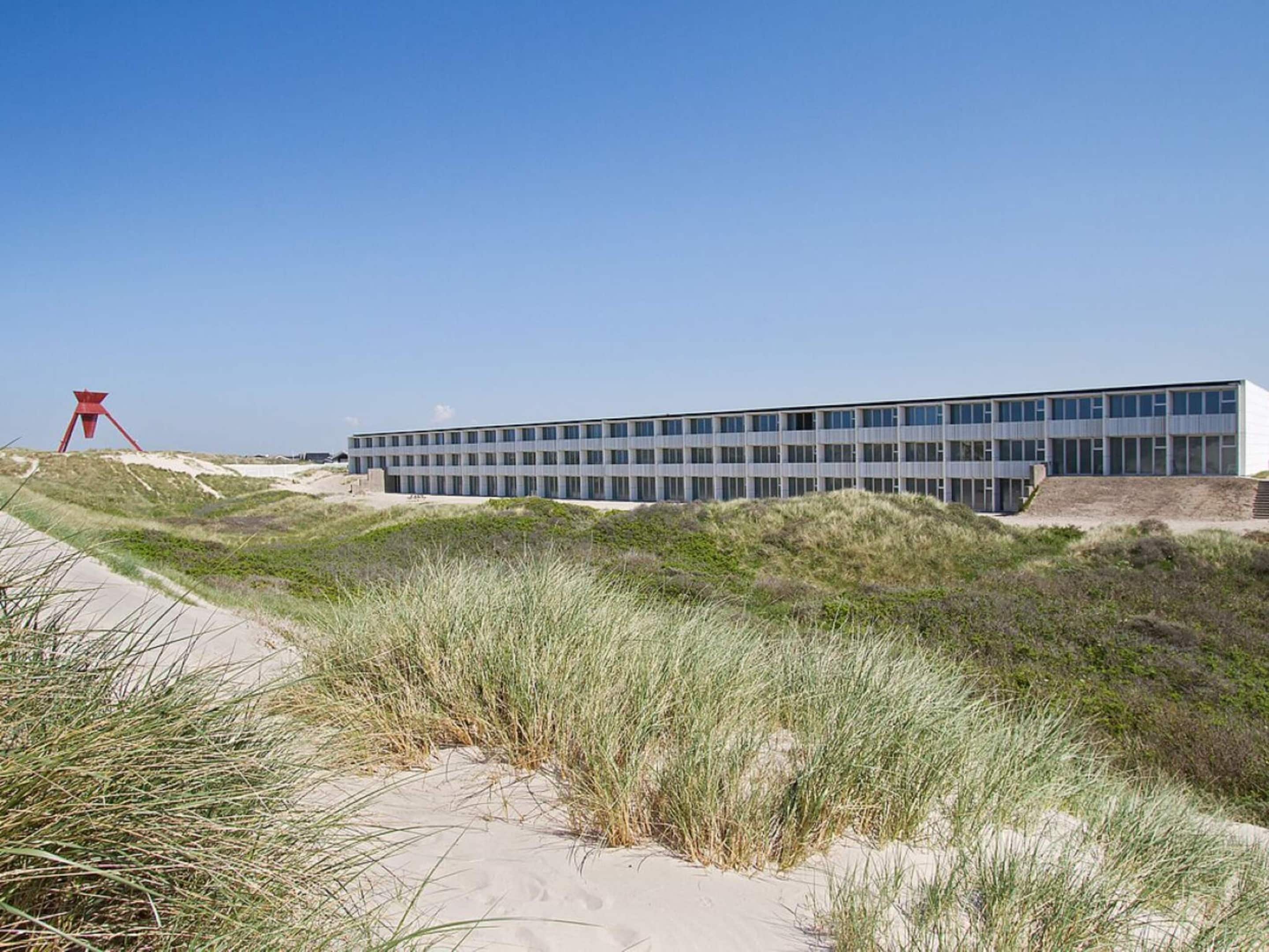 Un edificio moderno con techo plano y ventanas amplias, rodeado de dunas de arena y hierba.