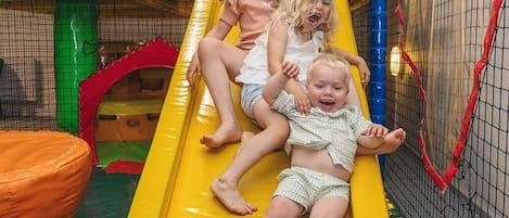 Children playing on a yellow slide in an indoor play area with a netted enclosure.