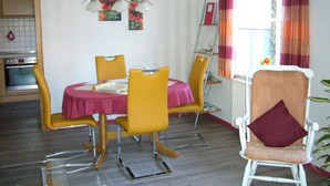 Un comedor con una mesa redonda, sillas amarillas y un mantel rojo. Se ve una ventana con cortinas naranjas y un radiador en la pared.
