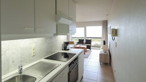 A modern kitchen with a built-in stove, sink, and dishwasher. There is a dining area with a table and chairs, and a window providing a view of the outdoors. - Apartment in Koksijde Near Sandy Beaches (Koksijde)