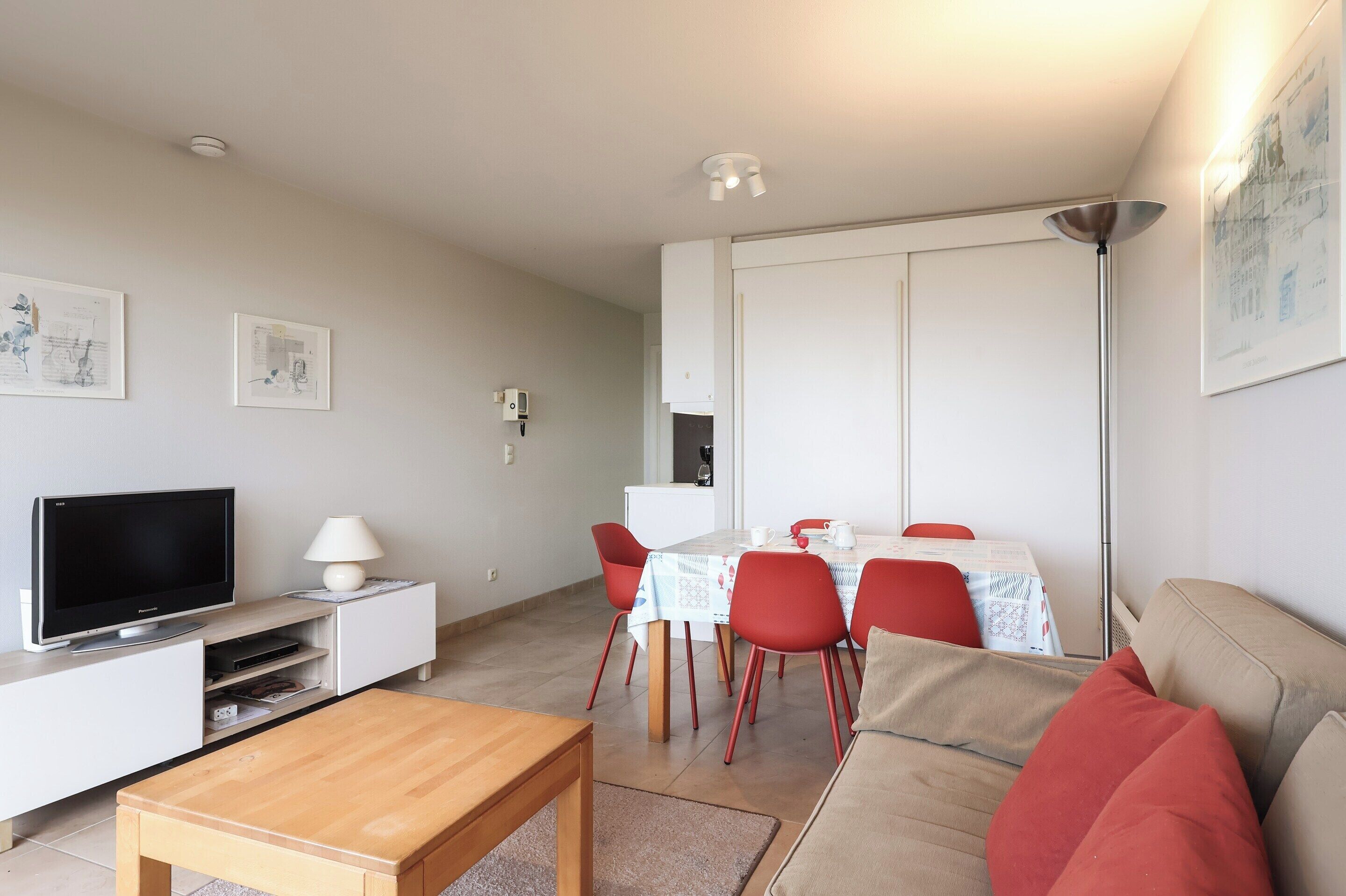 A modern living room with a flat-screen TV, a wooden coffee table, red chairs, a beige sofa, and a dining table with a white tablecloth.