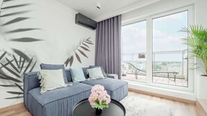 Ein modernes Wohnzimmer mit einem grauen Sofa, einem schwarzen Couchtisch mit einer rosa Blumenkomposition und einem großen Fenster mit Blick auf eine Stadtlandschaft.