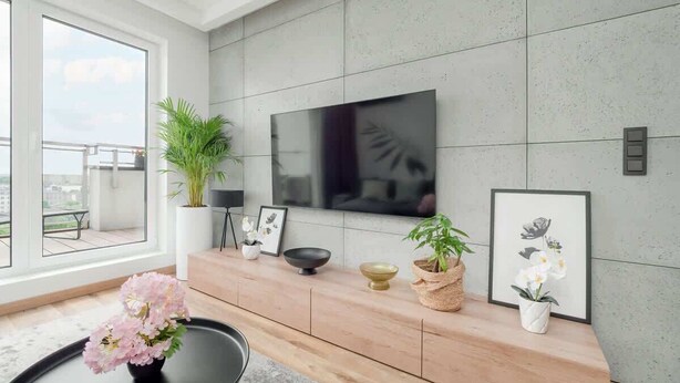 Ein modernes Wohnzimmer mit einem großen Flachbildfernseher an einer gefliesten Wand, einem Holzkonsolentisch mit Dekorationen und Blick auf einen Balkon durch große Fenster.